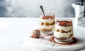 Dwa deserki tiramisu w szklankach, posypane kakao, na marmurowej desce.
