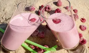 Dwa szklanki różowego koktajlu z malinami na piasku, obok zielone słomki i maliny.