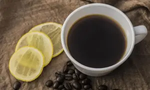 Biała filiżanka kawy z cytryną i ziarnami kawy na brązowym tle.