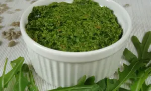 Zielone pesto z rukoli w białej miseczce, otoczone liśćmi rukoli i nasionami słonecznika.