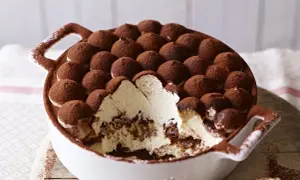 Tiramisu z mascarpone w białej misie, posypane kakao, z widocznym kremem i biszkoptami.