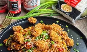 Talerz z makaronem udon, panierowanym kurczakiem, sezamem i szczypiorkiem.