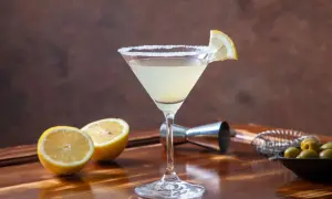 Koktajl Martini Bianco w kieliszku z solą na brzegu, ozdobiony plasterkiem cytryny, obok połówki cytryny i oliwek.
