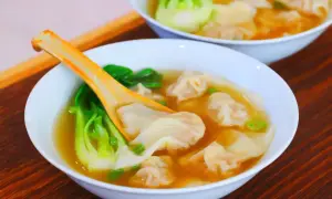 Dwie białe miski z zupą wonton, z widocznymi pierożkami, zielonymi warzywami i pomarańczową łyżką.