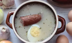 Miseczka żurku z kiełbasą i jajkiem, otoczona cebulą, czosnkiem i jajkami.