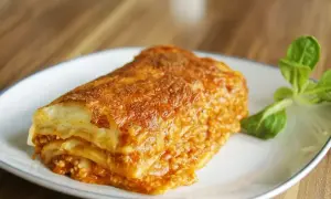 Kawałek zapieczonej lasagne z mięsem mielonym na białym talerzu, udekorowany listkami sałaty.