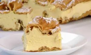 Kawałek karpatki z kremem na talerzyku, w tle całe ciasto posypane cukrem pudrem.