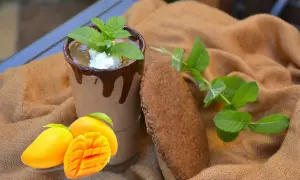 Szklanka mango lassi z czekoladą, miętą, mango i ciasteczkiem na brązowym tle.