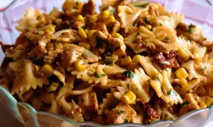 Sałatka z makaronem farfalle, kurczakiem, kukurydzą i suszonymi pomidorami w szklanej misie.