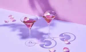 Dwa kieliszki z różowym drinkiem i płatkami róż na fioletowym tle.