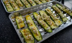 Plastry cukinii z serem feta i przyprawami na tacce aluminiowej, gotowe do grillowania.