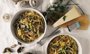 Dwa talerze makaronu z grzybami, posypane parmezanem i natką pietruszki. Obok świeże i suszone grzyby, kawałek parmezanu i tarka. Pyszny makaron z oliwą truflową.