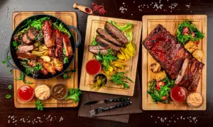 Trzy deski z grillowanym mięsem: żeberka, steki i skrzydełka kurczaka, podane z frytkami, kukurydzą i sosami.