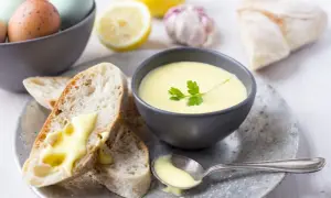 Kremowy sos aioli w miseczce, z pieczywem, jajkami, cytryną i czosnkiem w tle.