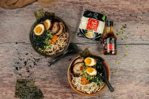 Dwie miski domowego ramenu z jajkiem, mięsem i nori, obok makaron i sos.