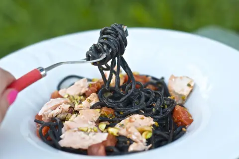 Czarny makaron z łososiem, pomidorkami i pistacjami na białym talerzu, podany na widelcu.