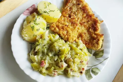 Kotlet schabowy z ziemniakami i zasmażaną kapustą z boczkiem na talerzu.