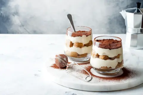 Dwa deserki tiramisu w szklankach, posypane kakao, na marmurowej desce.