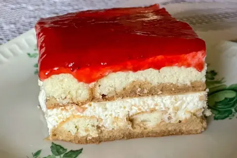 Kawałek ciasta bez pieczenia z warstwami biszkoptu, kremu i galaretki truskawkowej na talerzyku.