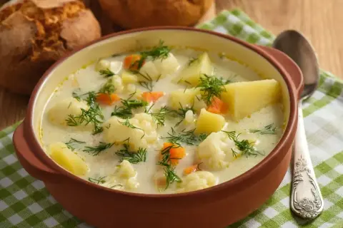 Miseczka kremowej zupy kalafiorowej z ziemniakami, marchewką i koperkiem, obok bułeczki.