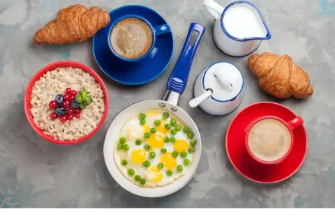 Jajecznica z groszkiem, owsianka z owocami, dwa croissanty i kawa na szarym tle.