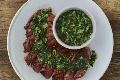 Pokrojony stek z wołowiny polany zielonym sosem chimichurri, podany na talerzu.