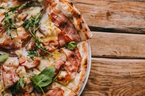 Kremowa pizza carbonara z boczkiem i rukolą na drewnianym stole.