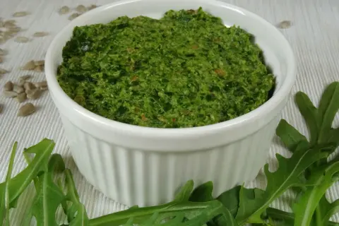 Zielone pesto z rukoli w białej miseczce, otoczone liśćmi rukoli i nasionami słonecznika.