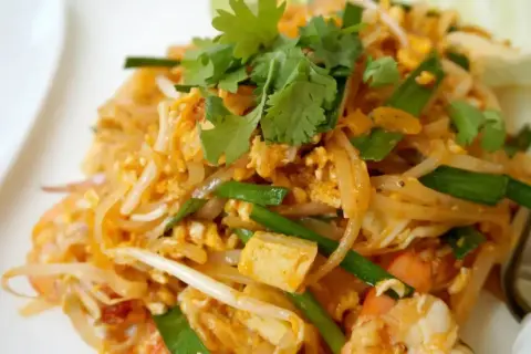 Zbliżenie na talerz z tajskim makaronem Pad Thai z krewetkami, jajkiem i szczypiorkiem, ozdobiony kolendrą.