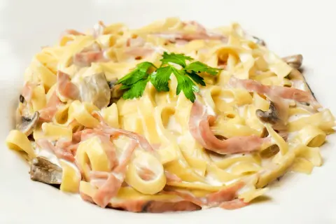 Makaron tagliatelle z szynką, pieczarkami i kremowym sosem, udekorowany natką pietruszki.