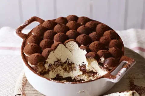 Tiramisu z mascarpone w białej misie, posypane kakao, z widocznym kremem i biszkoptami.