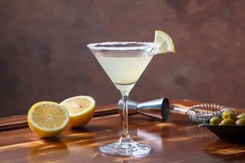 Koktajl Martini Bianco w kieliszku z solą na brzegu, ozdobiony plasterkiem cytryny, obok połówki cytryny i oliwek.