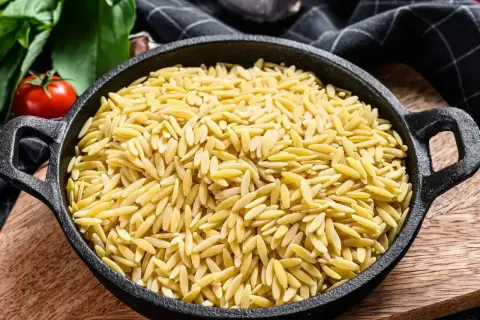 Makaron orzo w czarnej patelni, obok pomidorów i ziół.