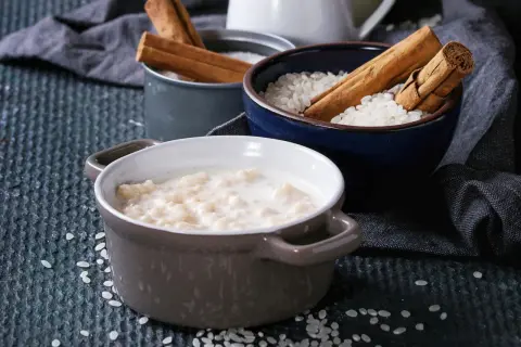 Gotowy ryż na mleku w miseczce, obok miseczki z ryżem i laskami cynamonu.
