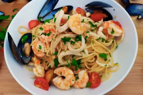 Talerz spaghetti z owocami morza: krewetkami, małżami i kalmarami, ozdobiony pomidorkami i natką pietruszki.