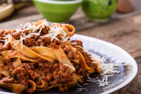 Talerz makaronu pappardelle z sosem bolognese i startym parmezanem.