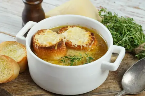 Aksamitna zupa cebulowa przepis, podana w białej miseczce, z grzankami zapieczonymi z serem i gałązką tymianku. Obok kromki chleba.