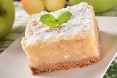 Kawałek ciasta z jabłkami, posypany cukrem pudrem i udekorowany listkiem mięty.