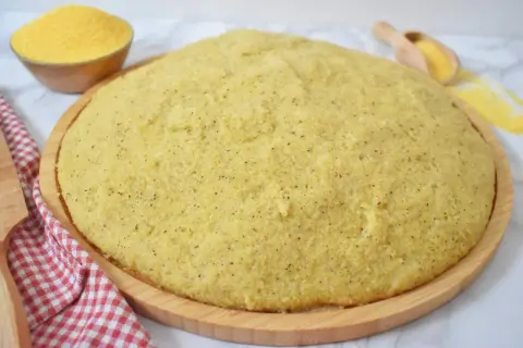 Gotowa, puszysta polenta na drewnianej desce, obok miseczka z mąką kukurydzianą.