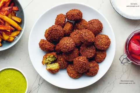 Talerz pełen złotobrązowych falafeli, jeden przekrojony, ukazujący zielone wnętrze. Obok frytki i sos.