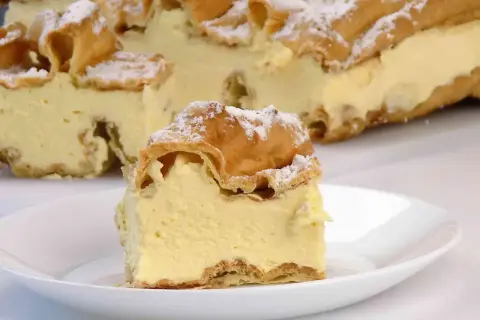 Kawałek karpatki z kremem na talerzyku, w tle całe ciasto posypane cukrem pudrem.