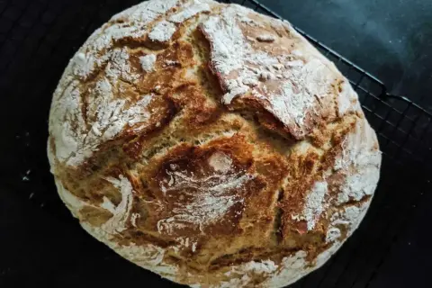 Złocisty, domowy bochenek chleba z chrupiącą skórką, posypany mąką, na czarnej kratce.
