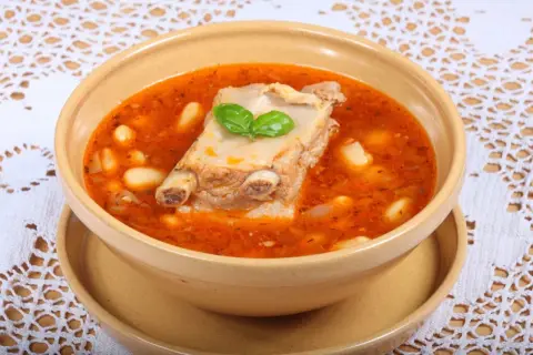 Aromaticzna zupa na żeberkach z fasolą i ziołami, podana w glinianej misce na ozdobnym obrusie.