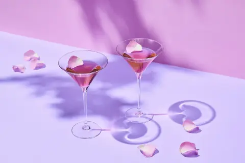 Dwa kieliszki z różowym drinkiem i płatkami róż na fioletowym tle.