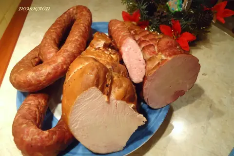 Wędzona szynka i kiełbasa na niebieskim talerzu, ozdobione gałązką choinki.