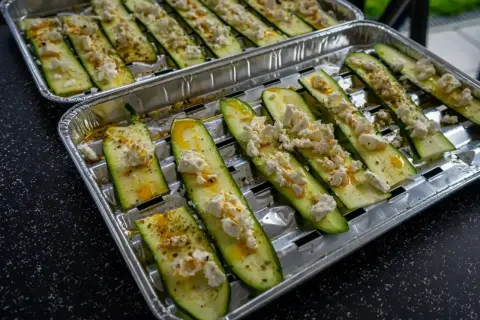 Plastry cukinii z serem feta i przyprawami na tacce aluminiowej, gotowe do grillowania.