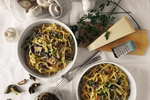 Dwa talerze makaronu z grzybami, posypane parmezanem i natką pietruszki. Obok świeże i suszone grzyby, kawałek parmezanu i tarka. Pyszny makaron z oliwą truflową.