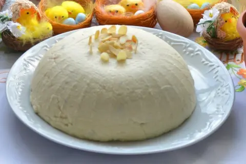 Pascha na białym talerzu, ozdobiona płatkami migdałów, w tle wielkanocne kurczaczki.