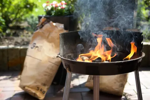Płonący grill z workami węgla drzewnego w tle.