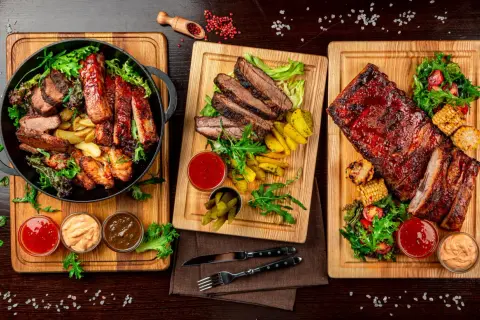 Trzy deski z grillowanym mięsem: żeberka, steki i skrzydełka kurczaka, podane z frytkami, kukurydzą i sosami.